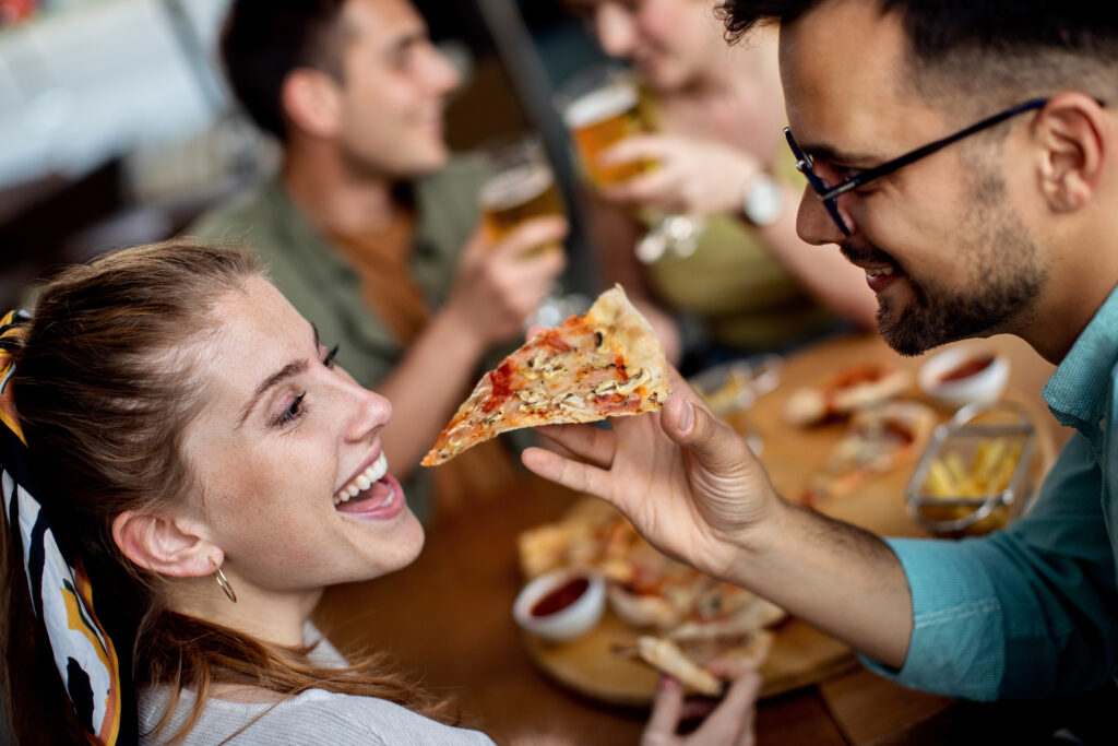 Happy man feeding his girlfriend with pizza while having lunch with friends in a pub.