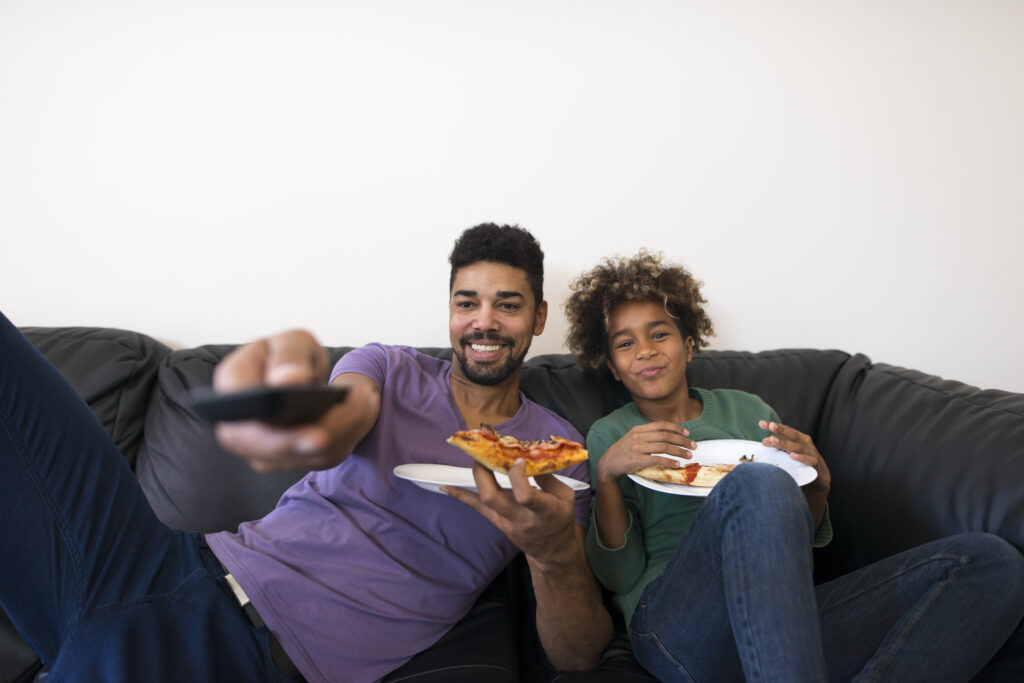 Happy father and daughter watching favorite TV show and enjoying pizza slice.